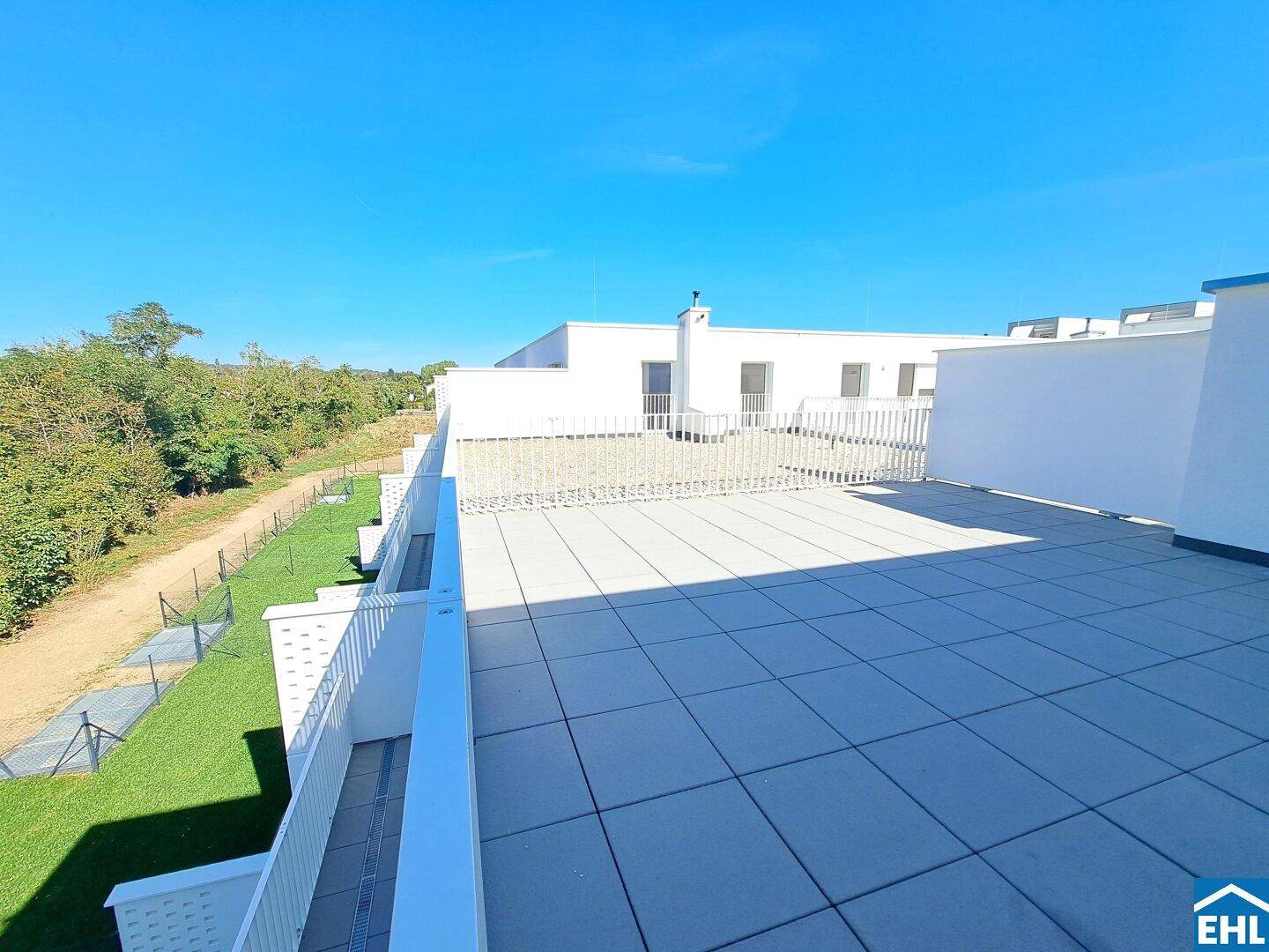 Weitläufige Dachterrasse mit Blick auf weitere Neubauten und grüne Umgebung unter blauem Himmel.
