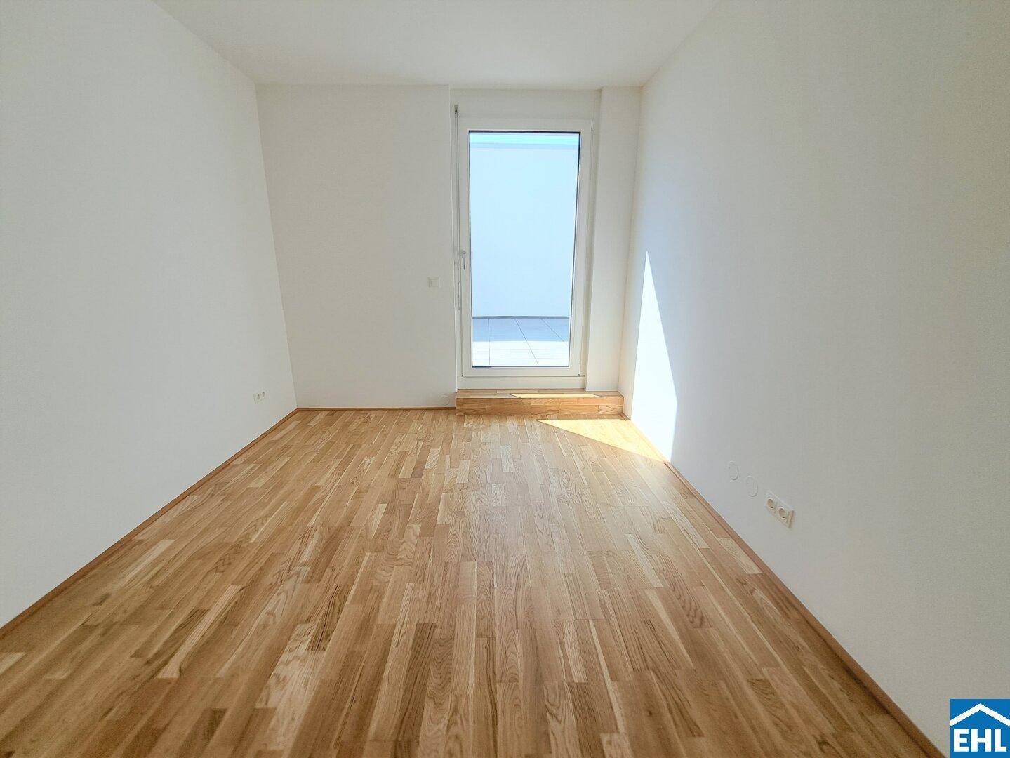 Helles, leeres Zimmer mit Parkettboden und Zugang zu einem Außenbereich.