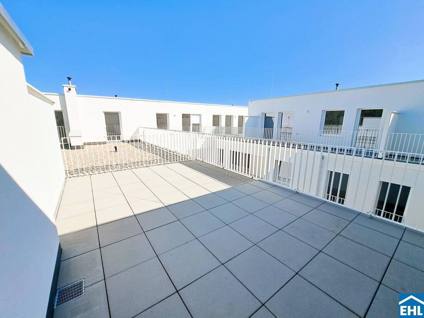 Weitläufige Dachterrasse mit Fliesenboden und weißem Geländer, umgeben von modernen Gebäuden.