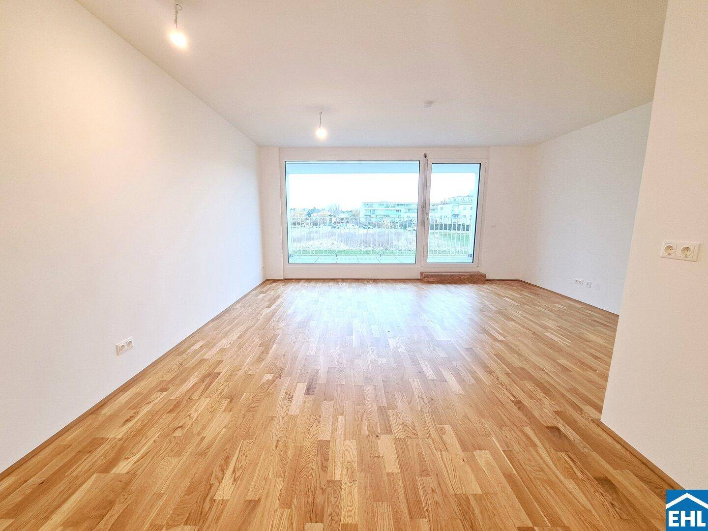 Heller, leerer Wohnraum mit großen Fenstern und neuem Parkettboden.