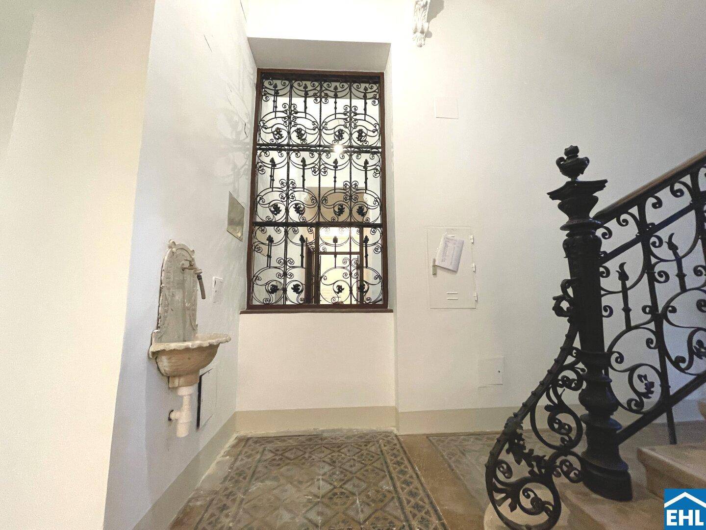 Historisches Treppenhaus mit schmiedeeisernem Geländer, großem Fenster und Steinwaschbecken.