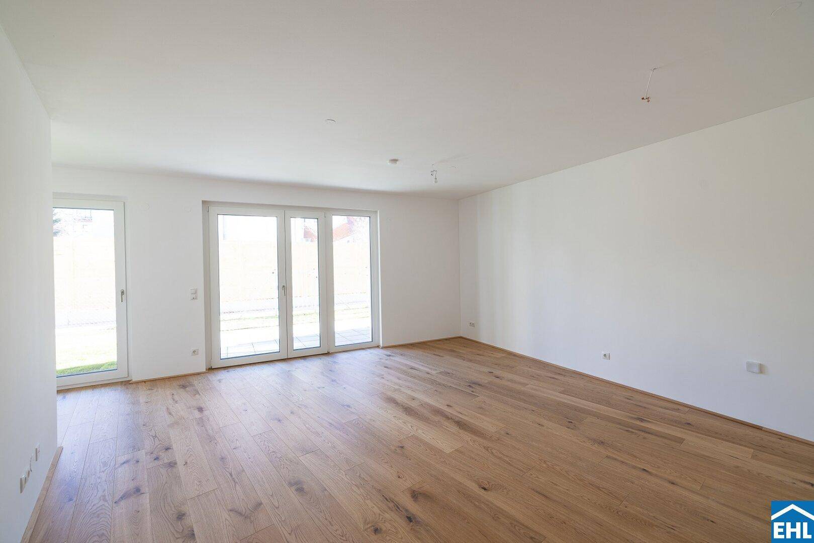 Geräumiger, leerer Raum mit hellem Holzboden und großen Fenstertüren zum Außenbereich.