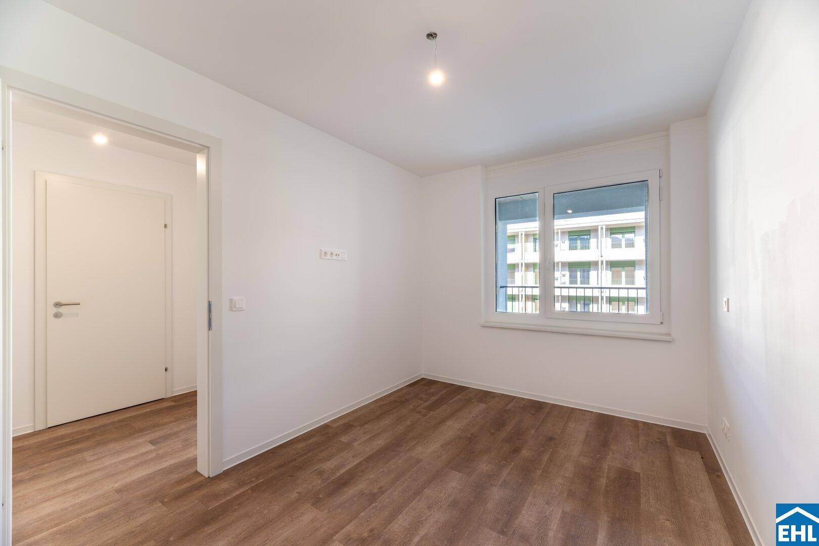 Helles Schlafzimmer mit Holzboden, weißen Wänden und Fensterblick auf die Nachbargebäude.