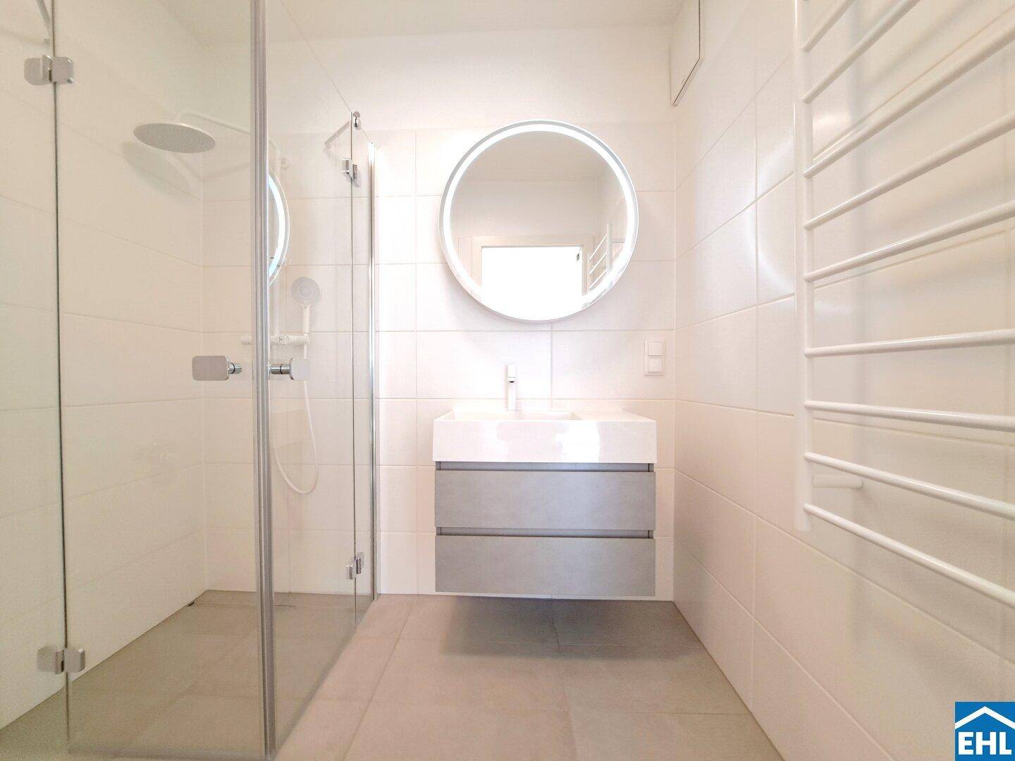 Modernes Badezimmer mit begehbarer Dusche, runden Spiegel und beheiztem Handtuchhalter.