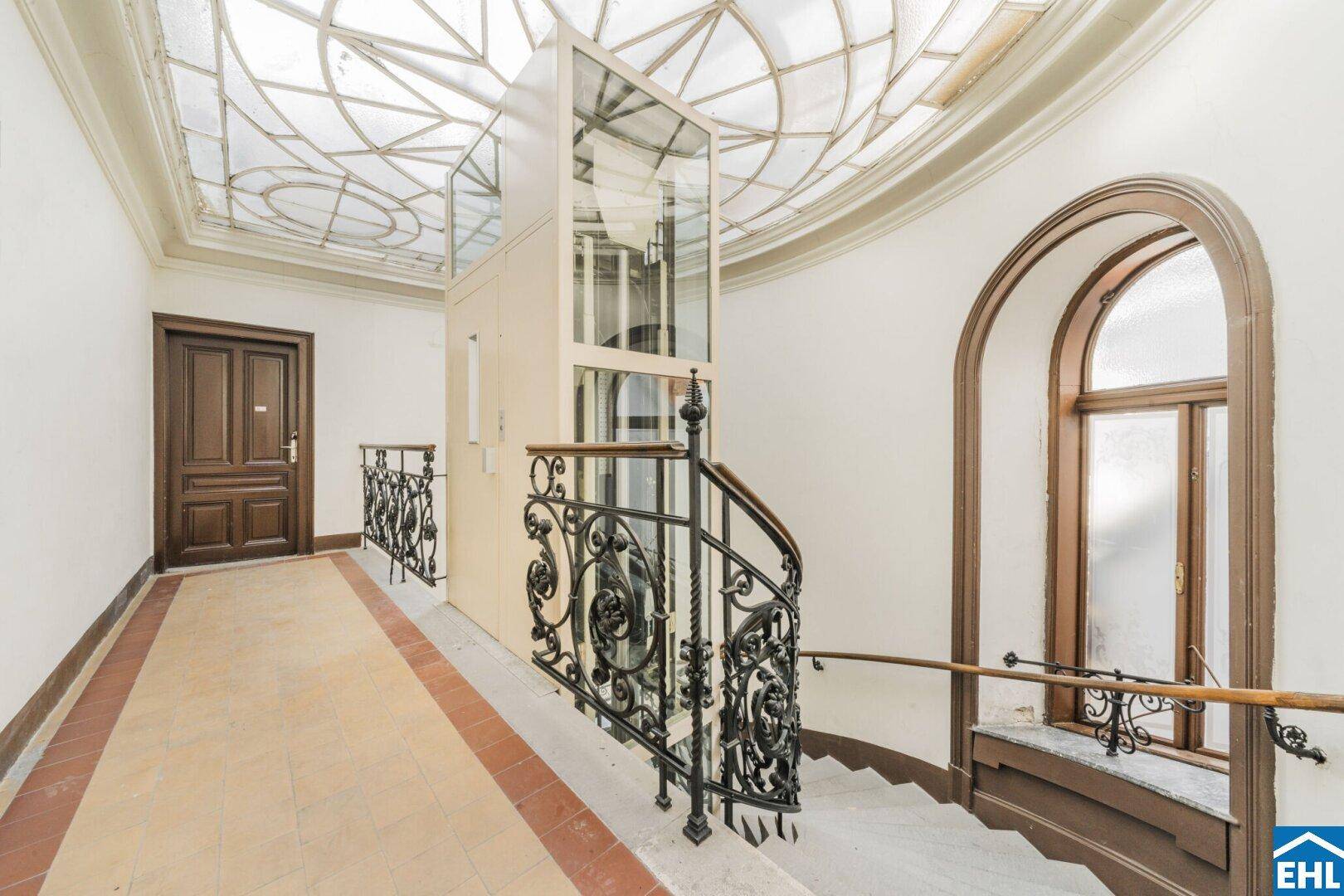 Helles Treppenhaus mit modernem Glasaufzug, schmiedeeisernem Geländer und dekorativer Deckenverglasung.