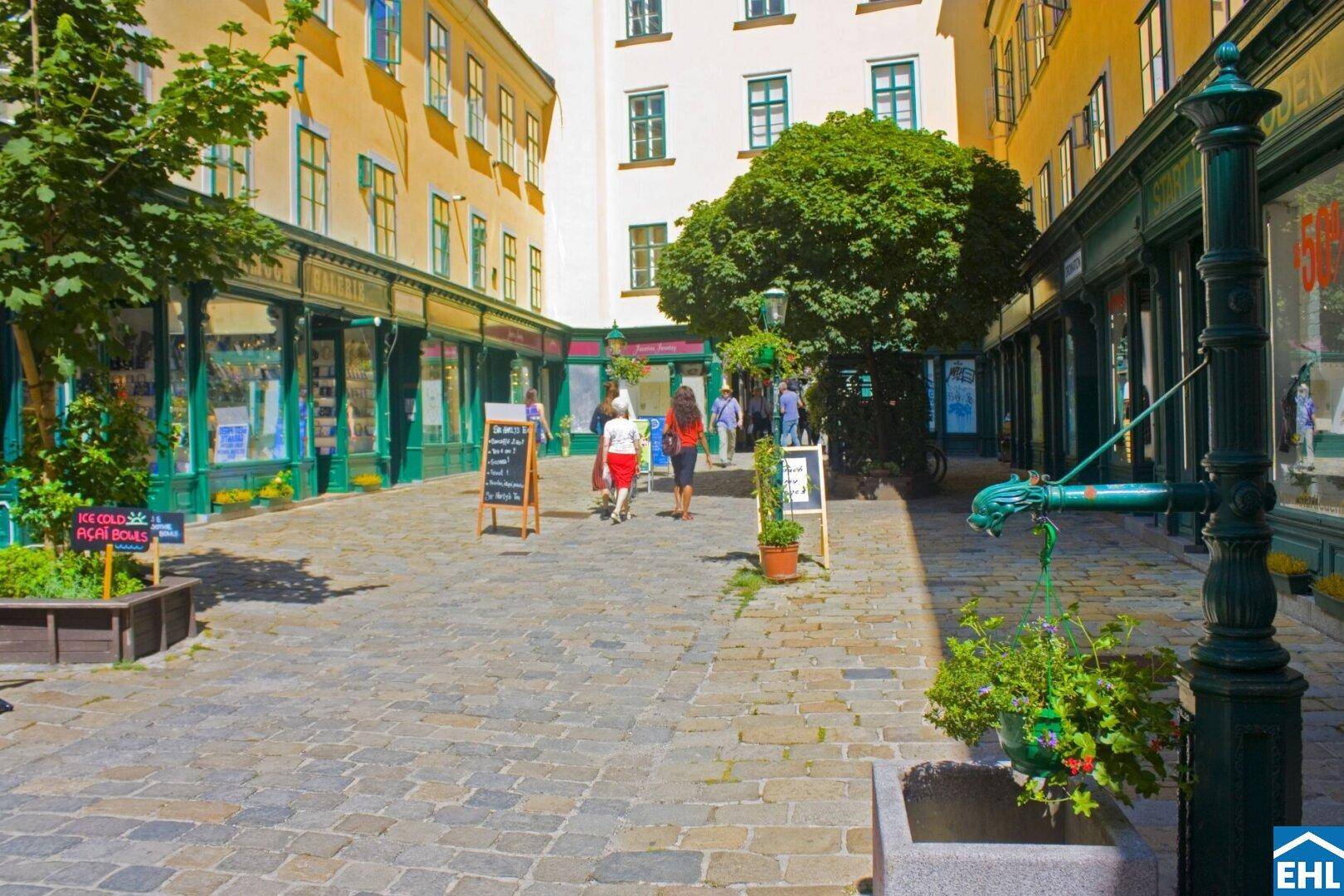 Belebter Innenhof mit kleinen Geschäften, Bäumen und einem historischen Brunnen an einem sonnigen Tag.