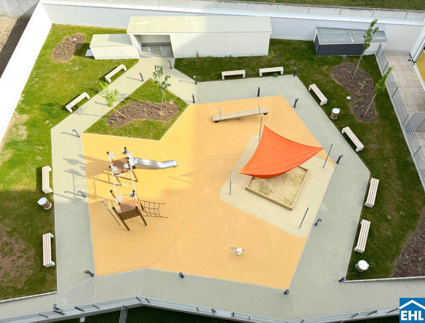 Blick von oben auf einen modernen Kinderspielplatz mit Sandkasten, Rutsche und Sitzbänken.