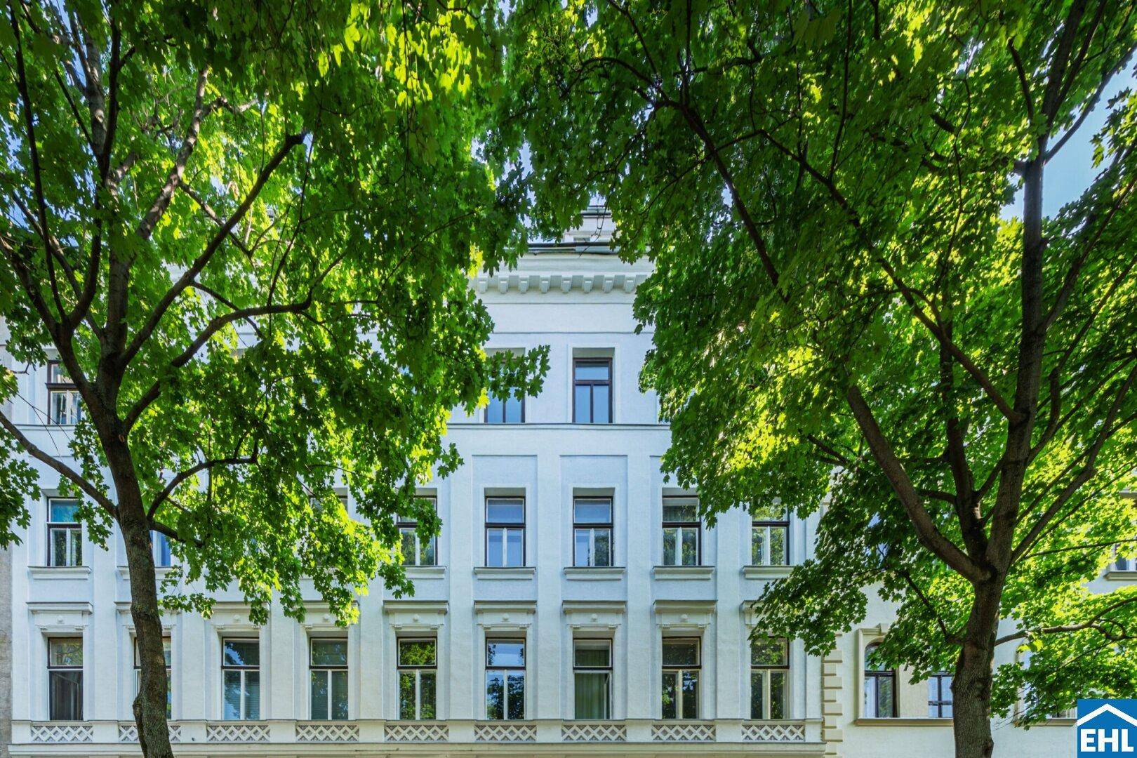 Klassische weiße Fassade eines Altbaus mit großen Fenstern, umrahmt von grünen Bäumen.