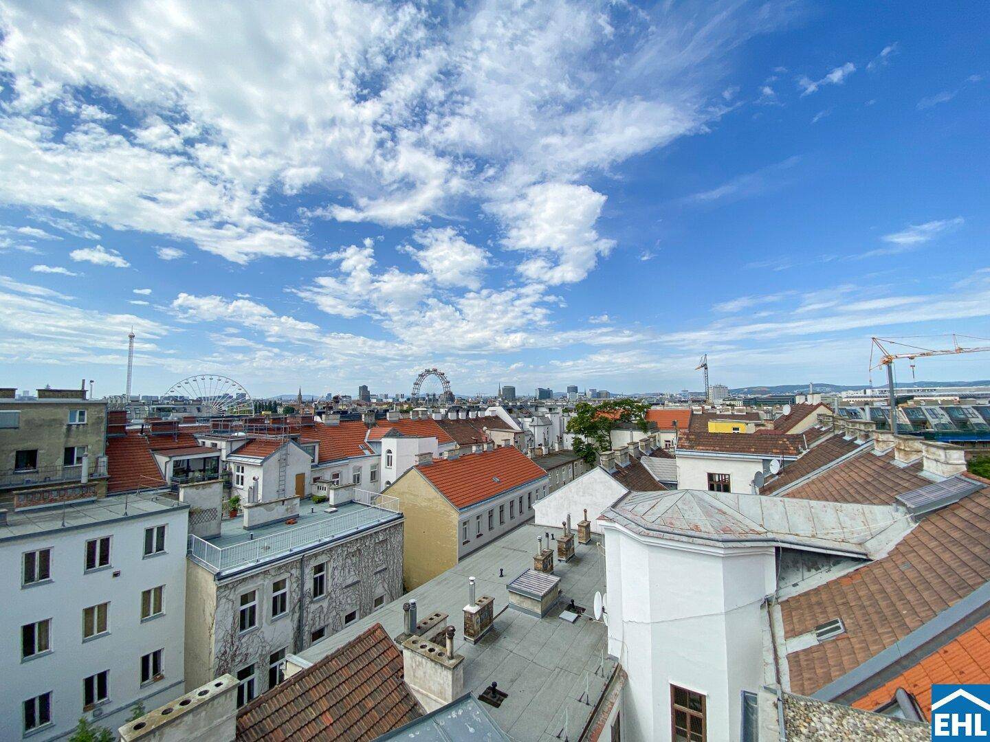 Weitläufiger Stadtblick über Dächer mit dem Wiener Riesenrad am Horizont unter einem blauen Himmel.