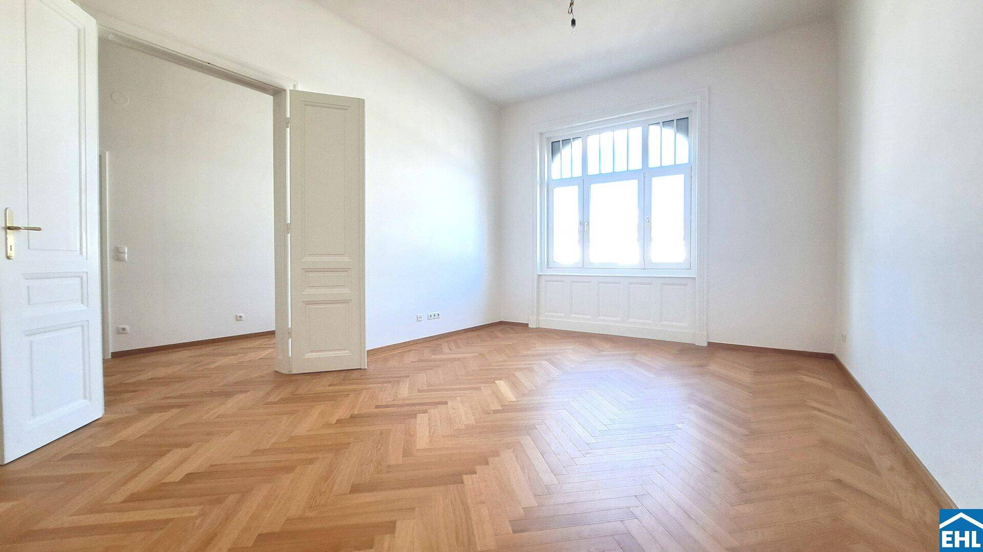 Helles Zimmer mit Fischgrätparkett und hohen Fenstern, das viel Tageslicht hereinlässt.
