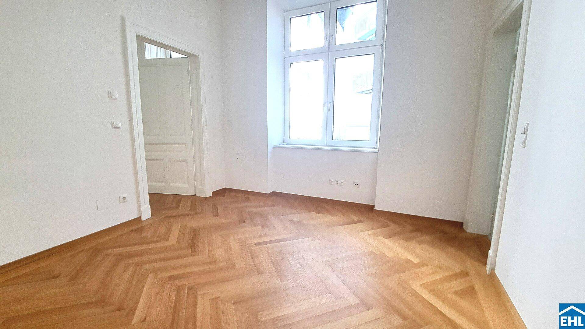 Lichtdurchfluteter Raum mit elegantem Fischgrätparkett und hohen Fenstern, bereit zur individuellen Gestaltung.