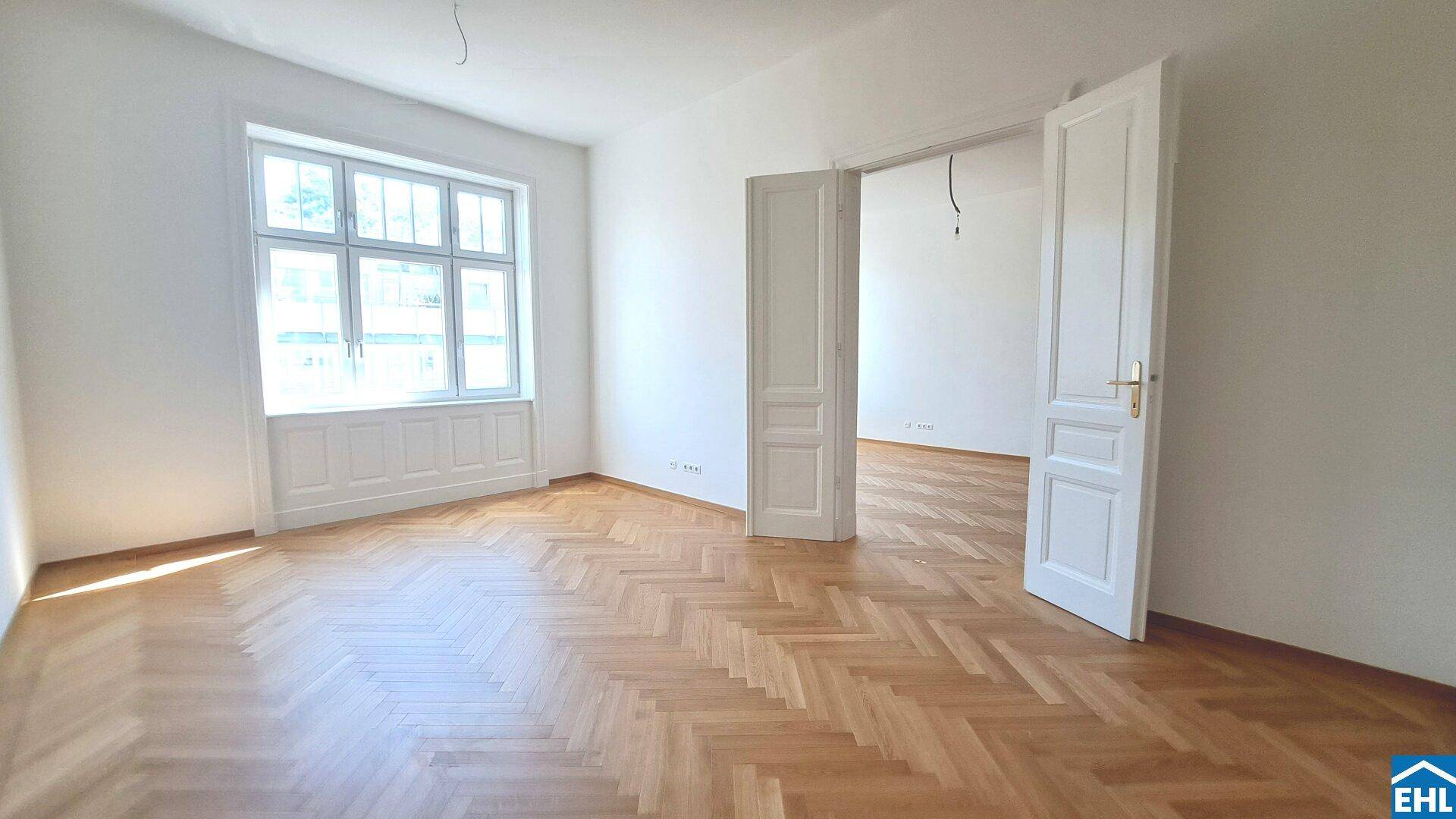 Geräumiges Wohnzimmer mit Fischgrätparkett und großem Fenster, das viel Tageslicht hereinlässt.