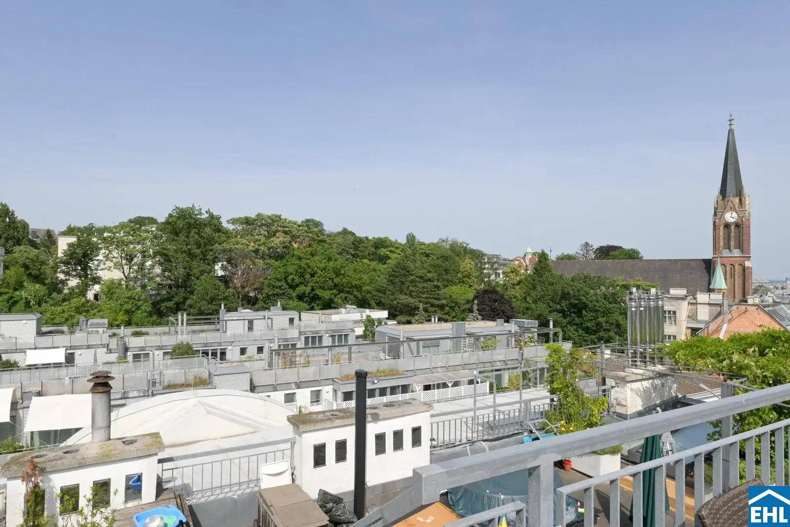 Weitläufiger Blick von der Dachterrasse über die umliegenden Gebäude, Bäume und eine markante Kirche.