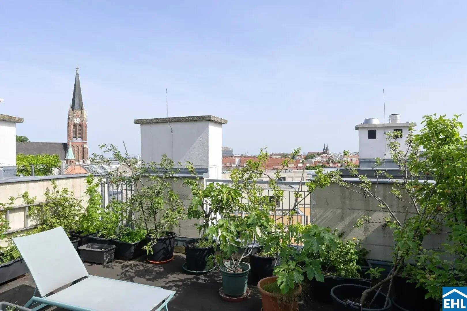 Sonnige Dachterrasse mit üppiger Bepflanzung und weitem Blick auf die Stadt und eine Kirchturmspitze.