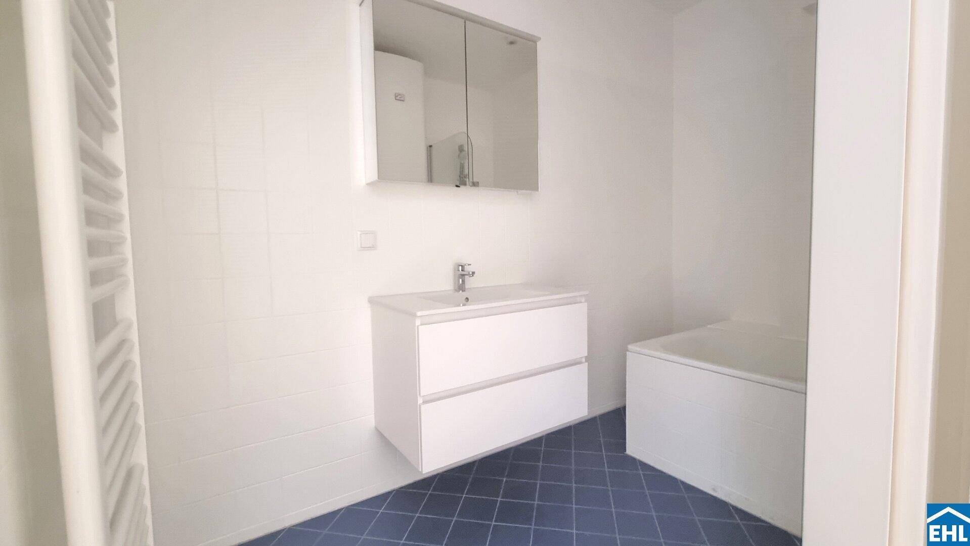 Modernes Badezimmer mit blauen Bodenfliesen, weißem Waschtisch und Badewanne sowie beheiztem Handtuchhalter.