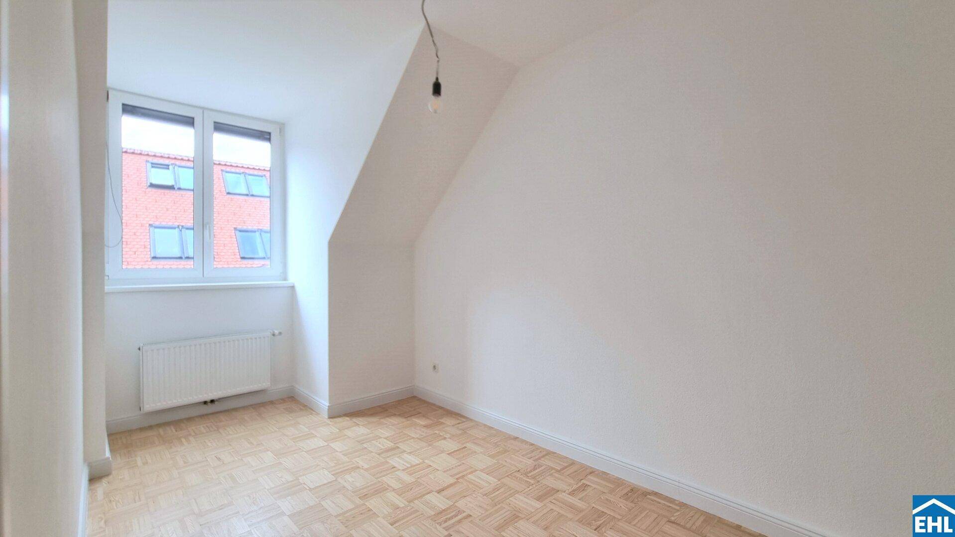 Helles Dachzimmer mit Parkettboden, weißen Wänden und einem Fenster, das viel Tageslicht hereinlässt.