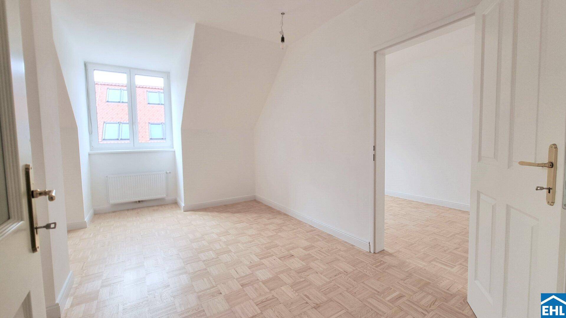 Helles Zimmer im Dachgeschoss mit Parkettboden, weiß gestrichenen Wänden und Fensterblick.