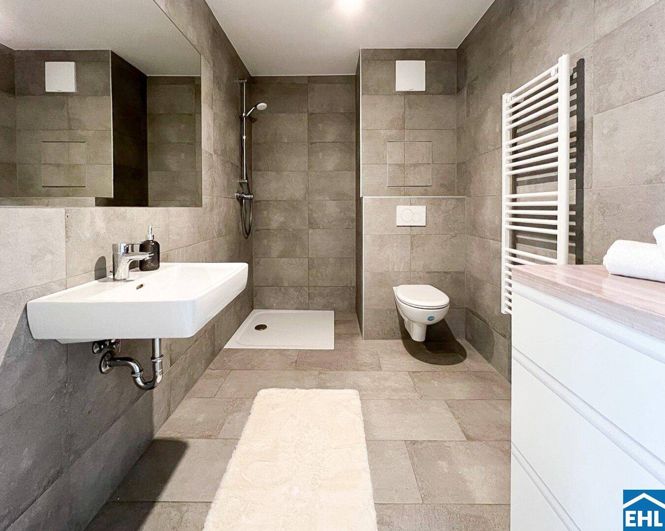 Modernes Badezimmer mit grauen Fliesen, ebenerdiger Dusche und beheiztem Handtuchhalter.