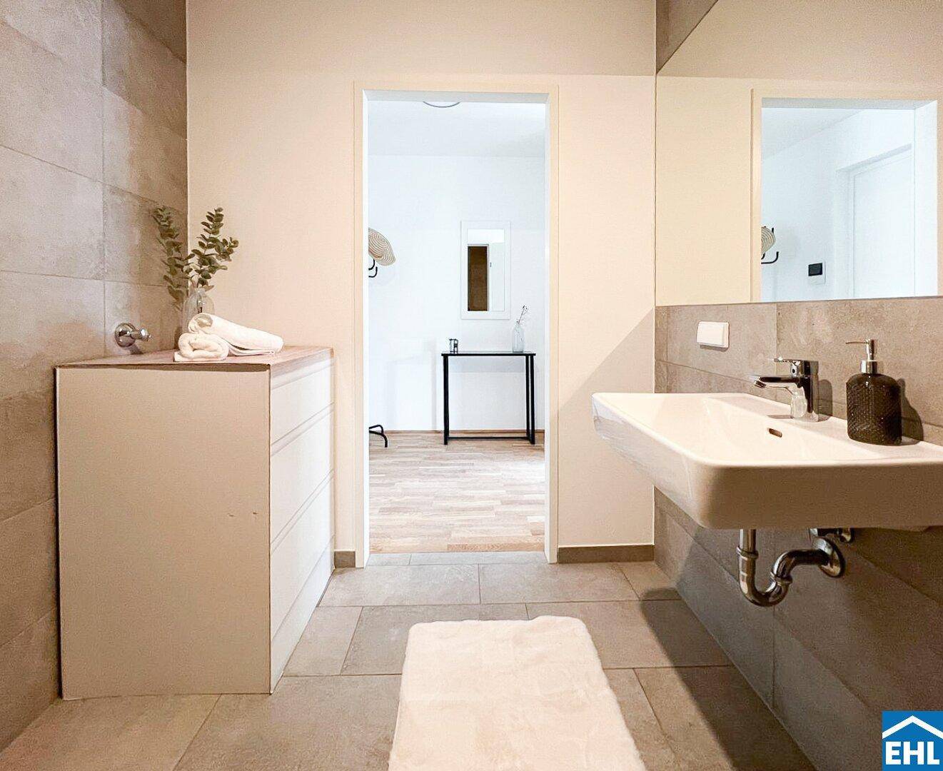Modernes Badezimmer mit Waschbecken, großem Spiegel und hellen Fliesen, das einen Blick in den Flur bietet.