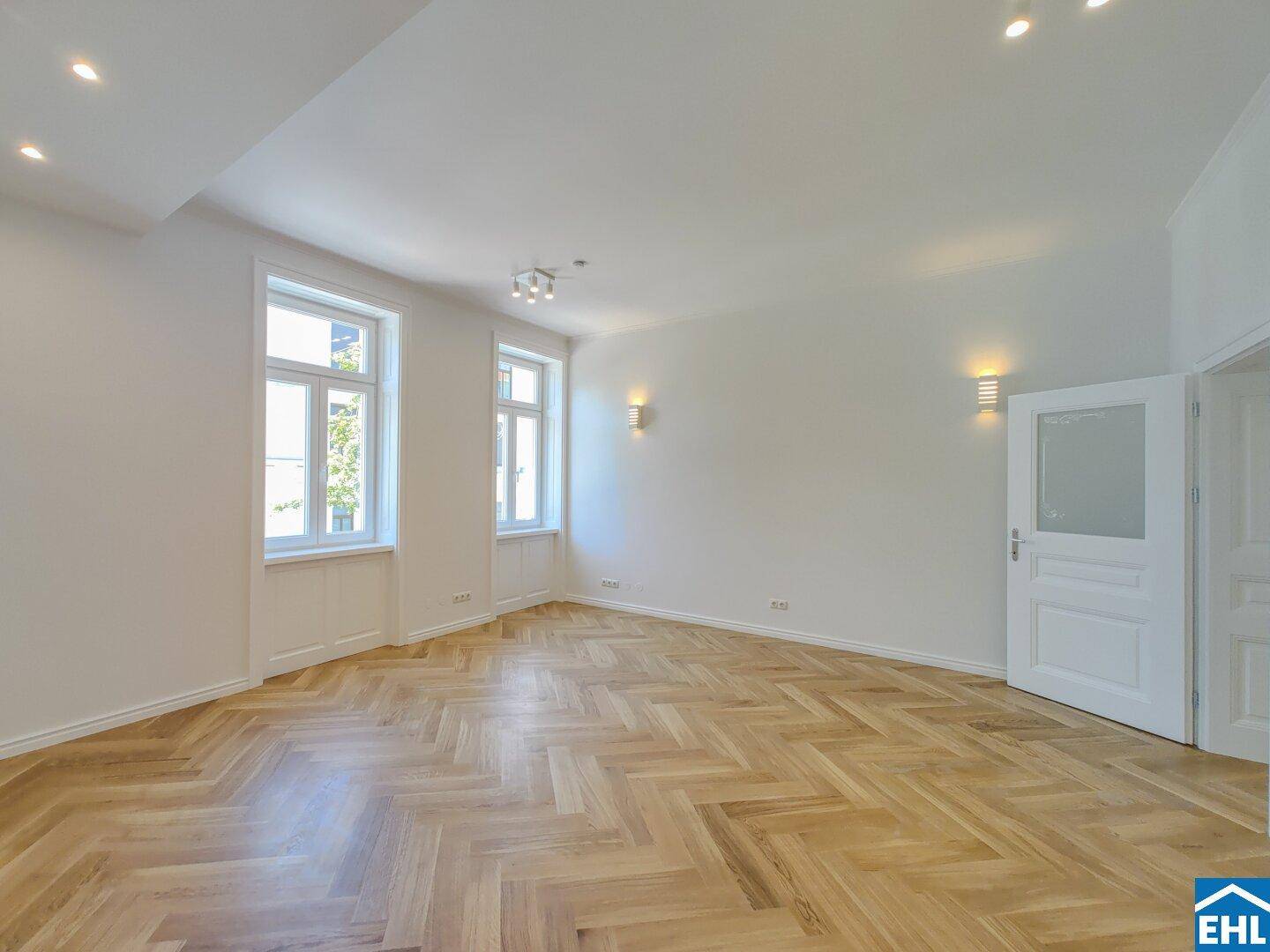 Helles Wohnzimmer mit Fischgrätparkett und hohen Fenstern, bereit für die individuelle Einrichtung.