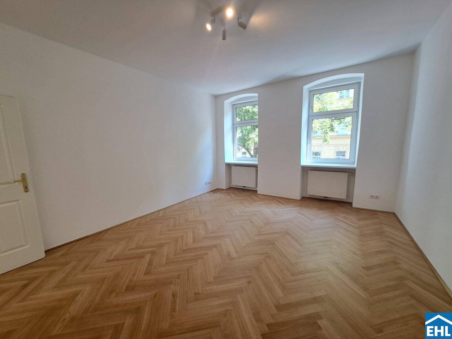 Lichtdurchflutetes Zimmer mit klassischem Fischgrätparkett und zwei Fenstern mit Blick ins Grüne.