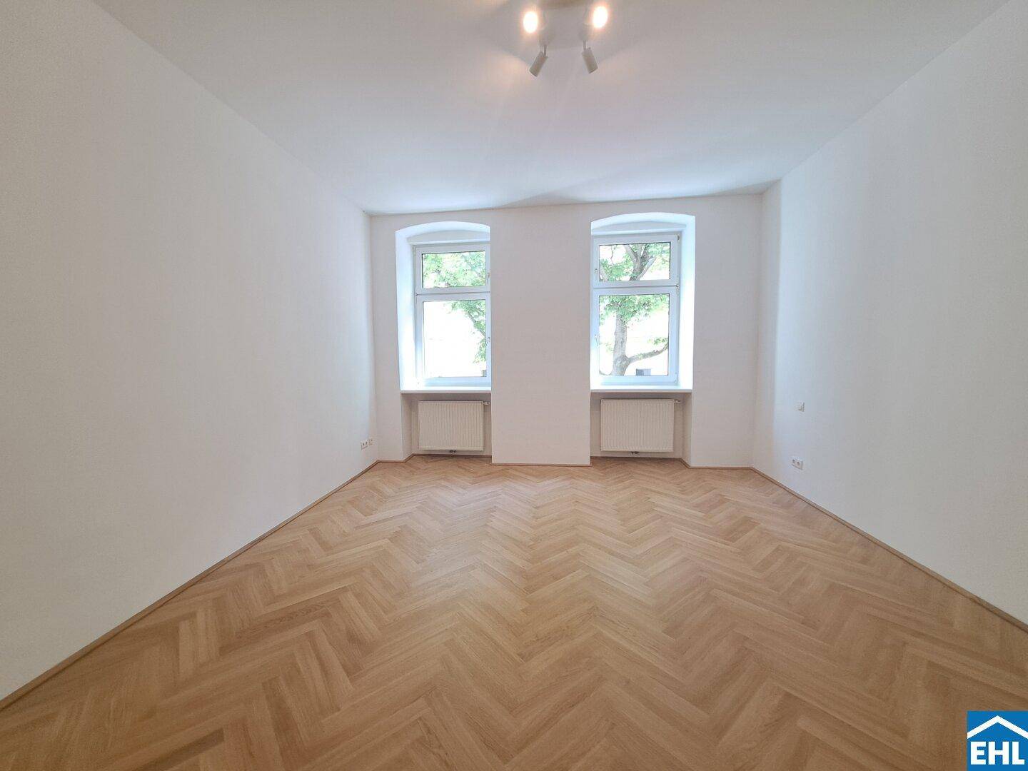 Helles Zimmer mit Fischgrätparkett und zwei Fenstern, das viel Tageslicht hereinlässt.