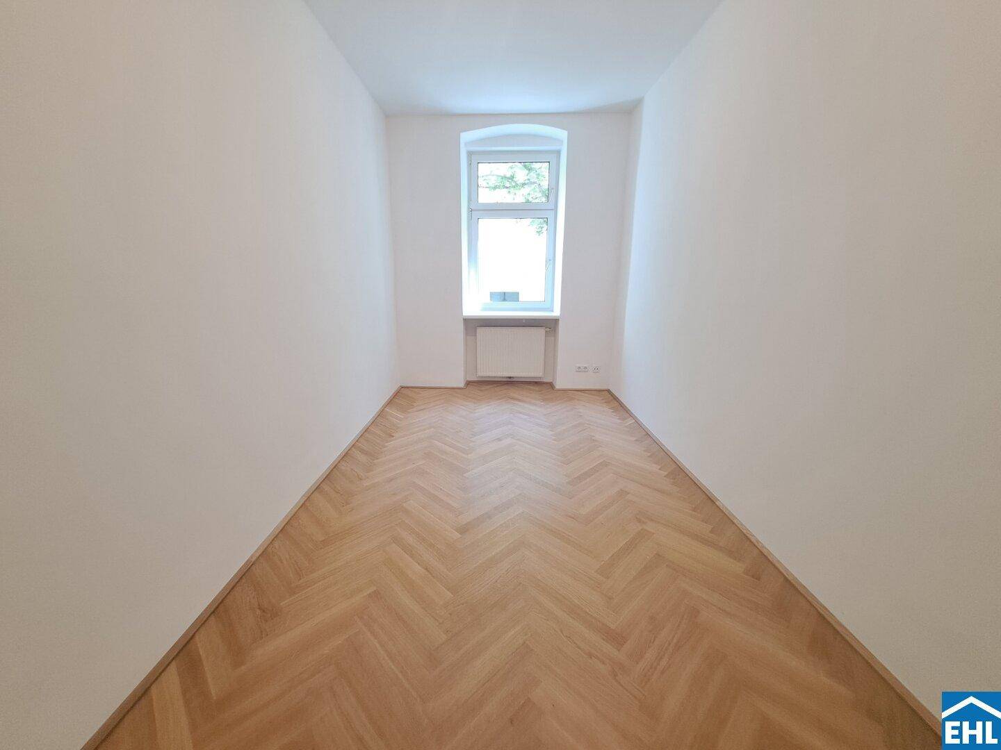 Langes, helles Zimmer mit Fischgrätparkett und einem einzelnen Fenster, das viel Tageslicht hereinlässt.