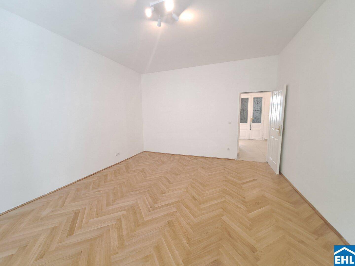 Großes, leeres Zimmer mit Fischgrätparkett und einer geöffneten Tür, die zu weiteren Räumen führt.