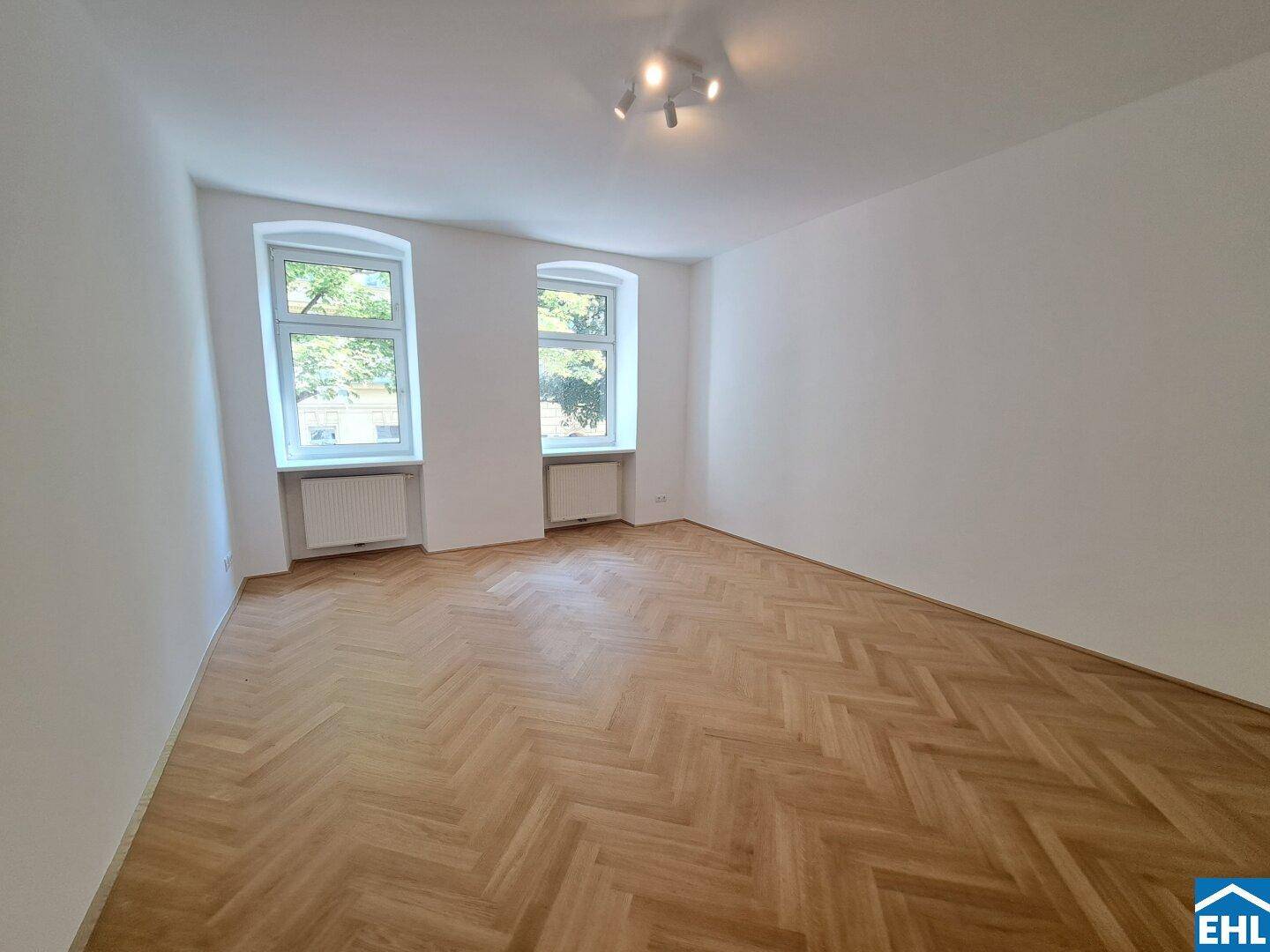Helles Zimmer mit Fischgrätparkett und zwei Fenstern, das viel Tageslicht hereinlässt.