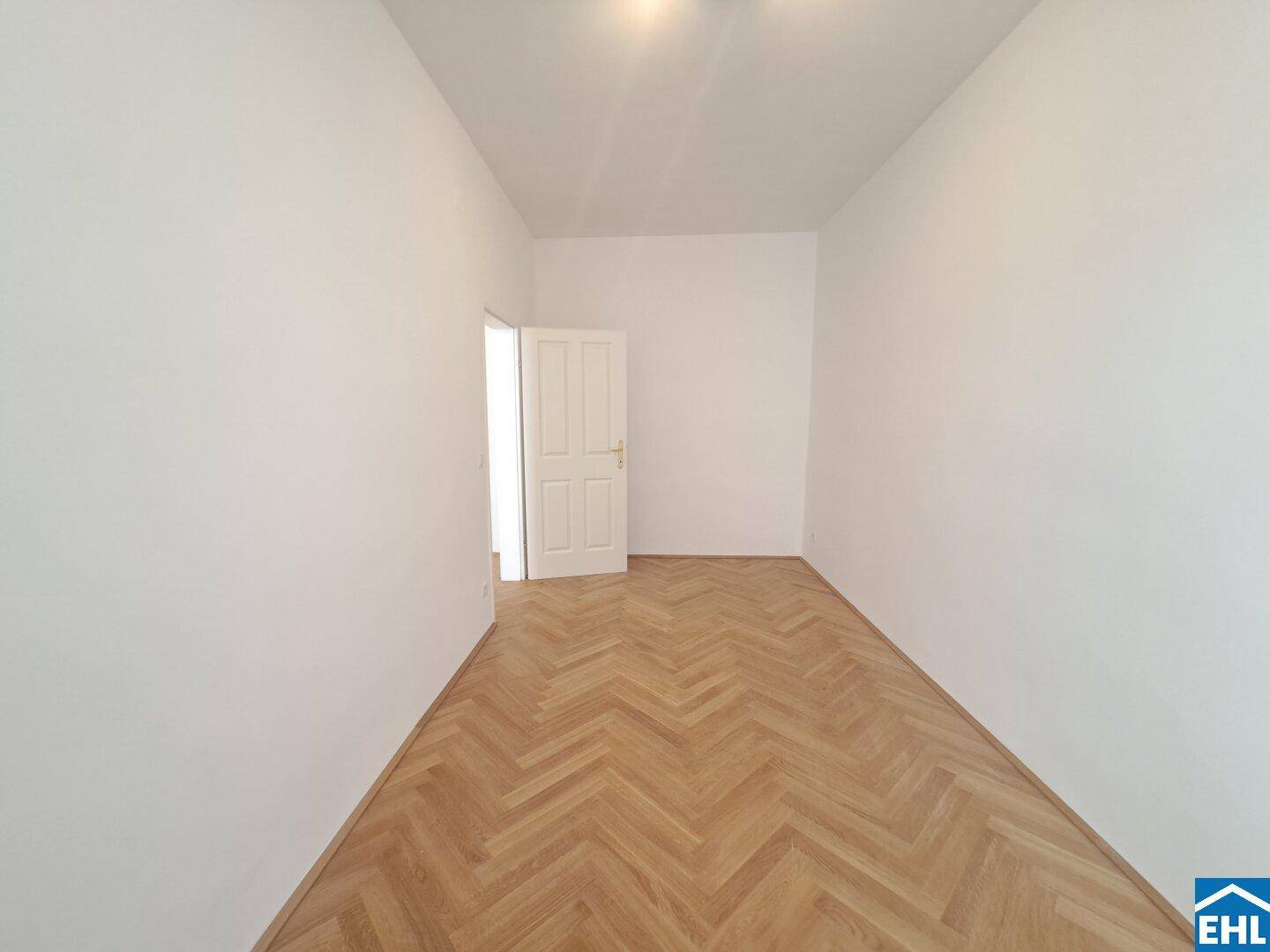 Langes, helles Zimmer mit Fischgrätparkett und einer geöffneten Tür, die den Blick freigibt.