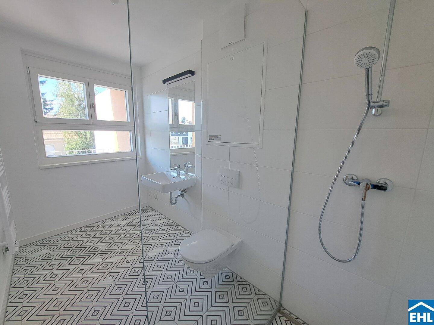 Helles Badezimmer mit Fenster, modernen Armaturen und auffälligem Fliesenbodenmuster im Duschbereich.