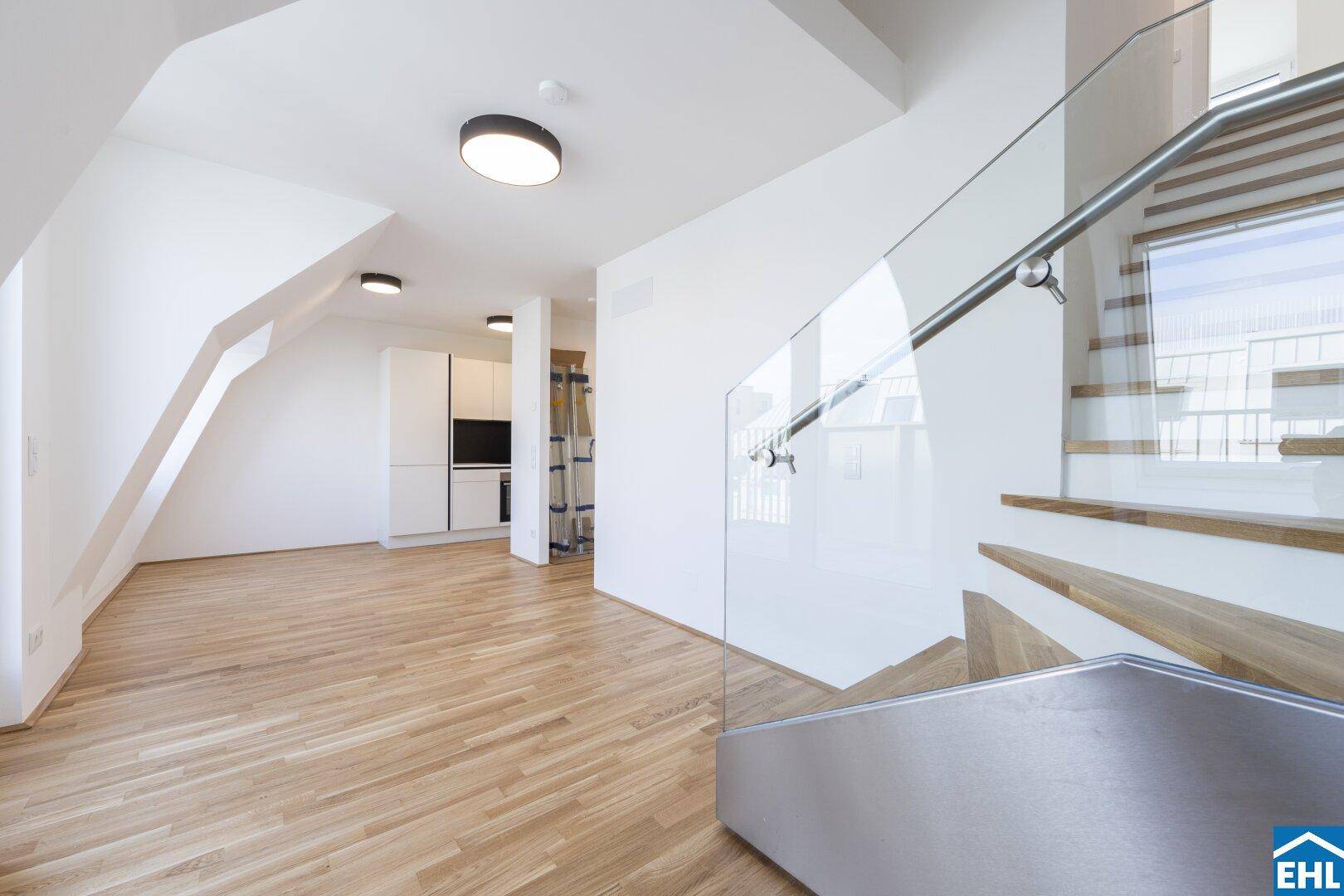 Moderner Wohnbereich mit Holzparkett, schräger Decke und eleganter Glastreppe, die zum oberen Stockwerk führt.