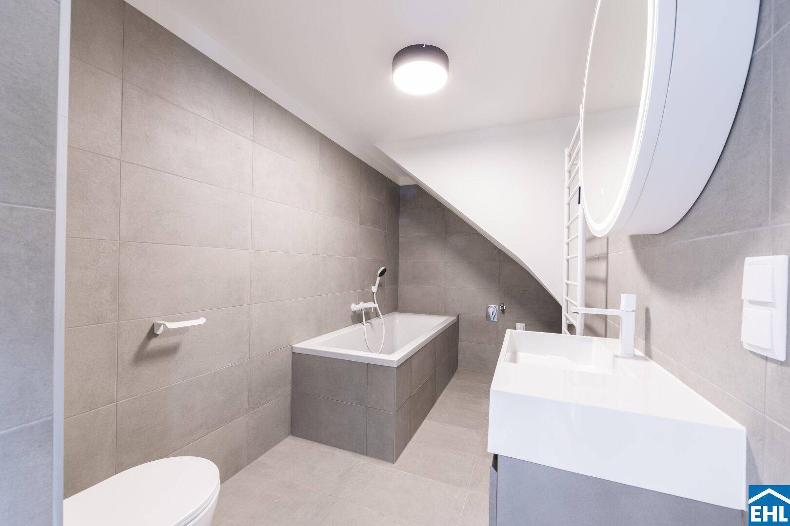 Modernes Badezimmer mit grauen Großformatfliesen, Badewanne, Waschtisch und schräger Decke.