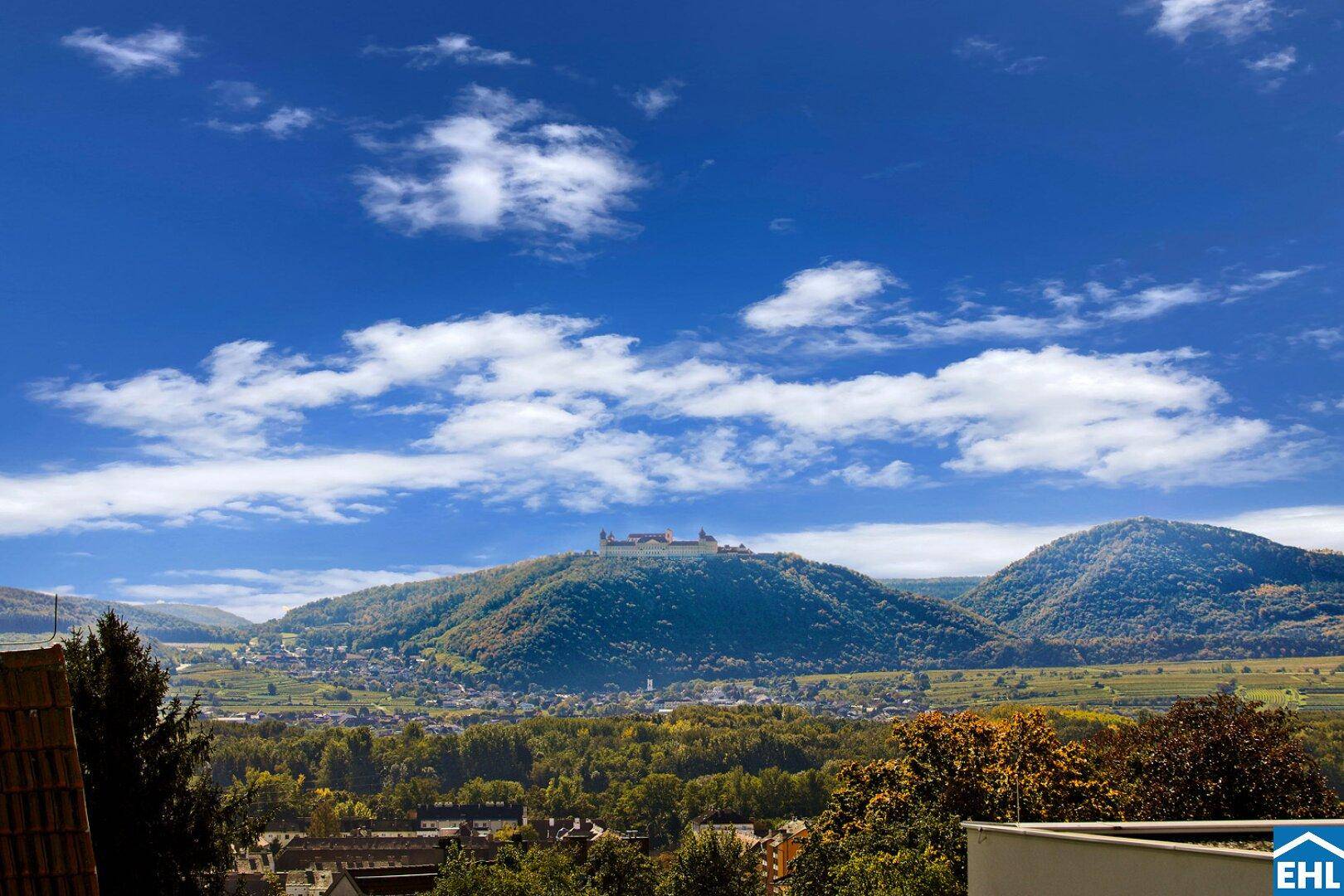 Panoramablick auf eine Burg auf einem Hügel, umgeben von grüner Landschaft und einem Dorf.