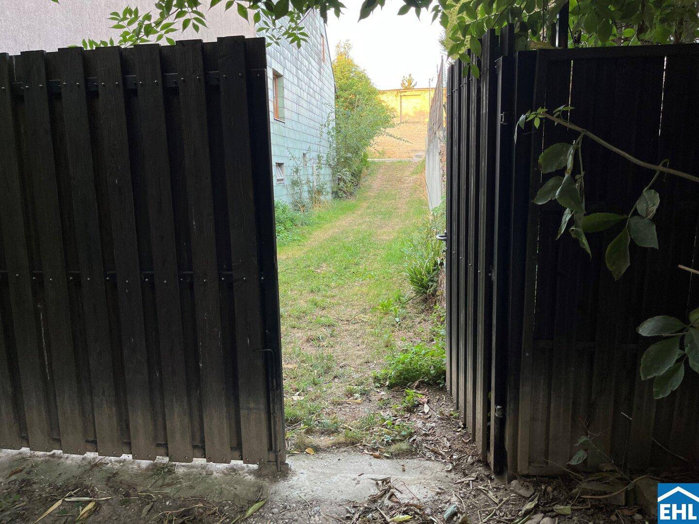 Offenes Holztor führt zu einem grasbewachsenen Weg, der zu weiteren Gebäuden in der Ferne führt.