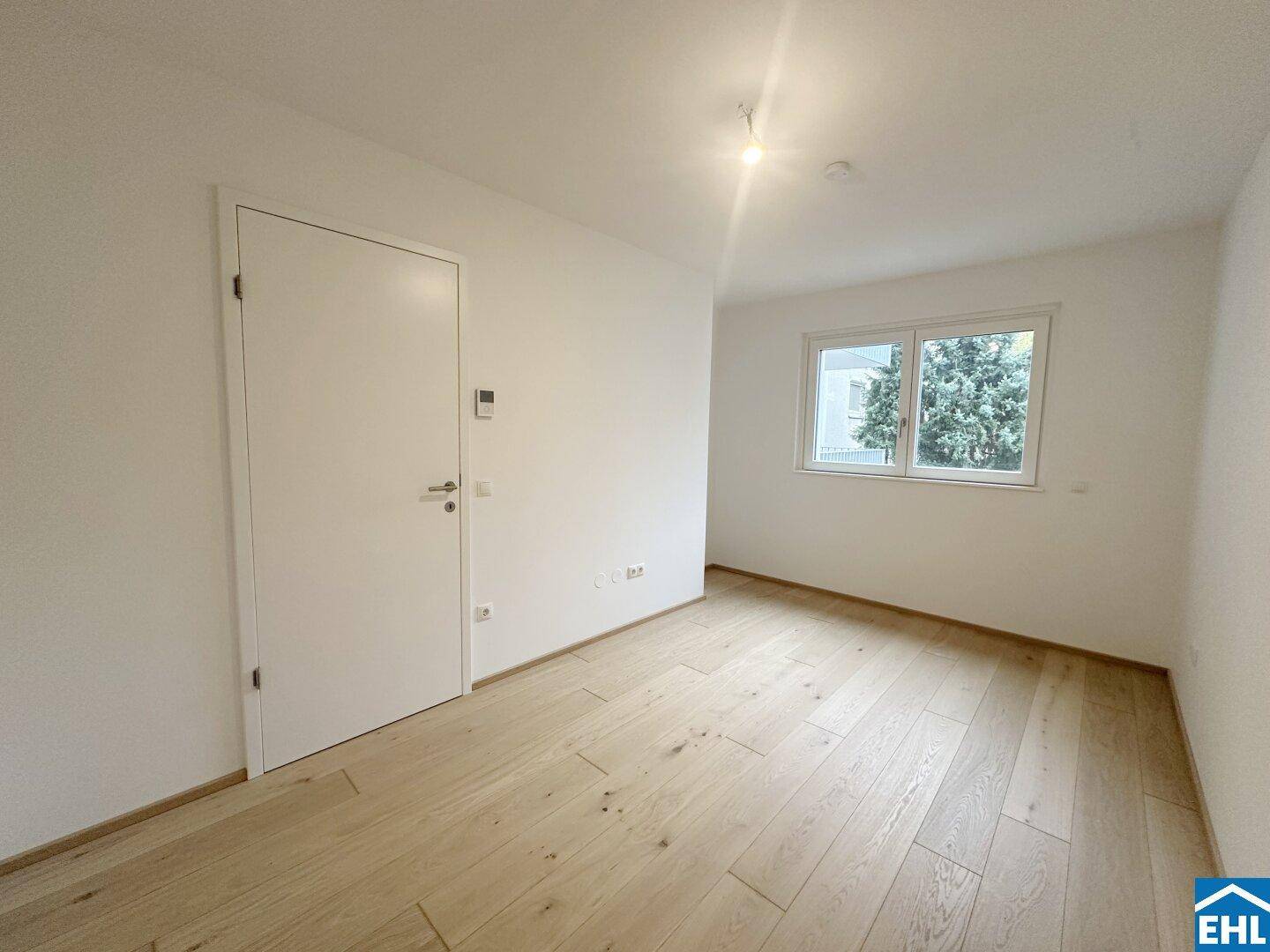 Ruhiges, unmöbliertes Zimmer mit Parkettboden und einem Fenster mit Blick ins Grüne.