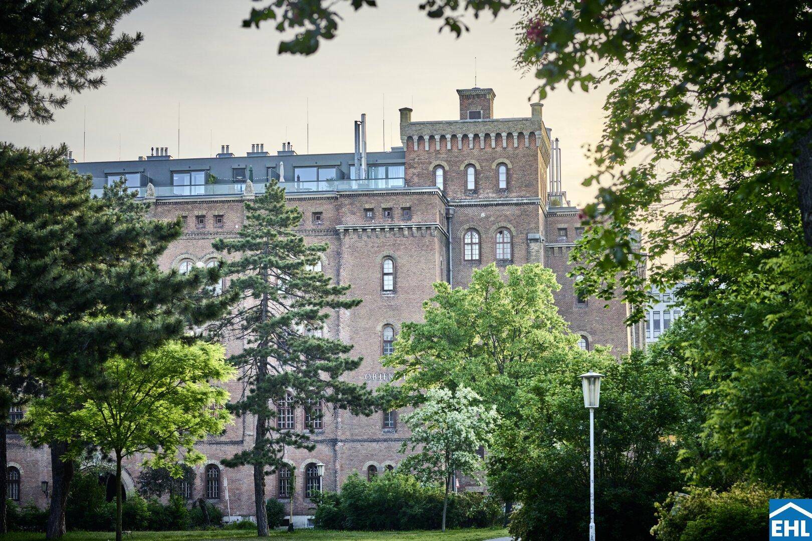 Historisches Backsteingebäude mit modernen Dachaufbauten, umgeben von Bäumen und Grünflächen.