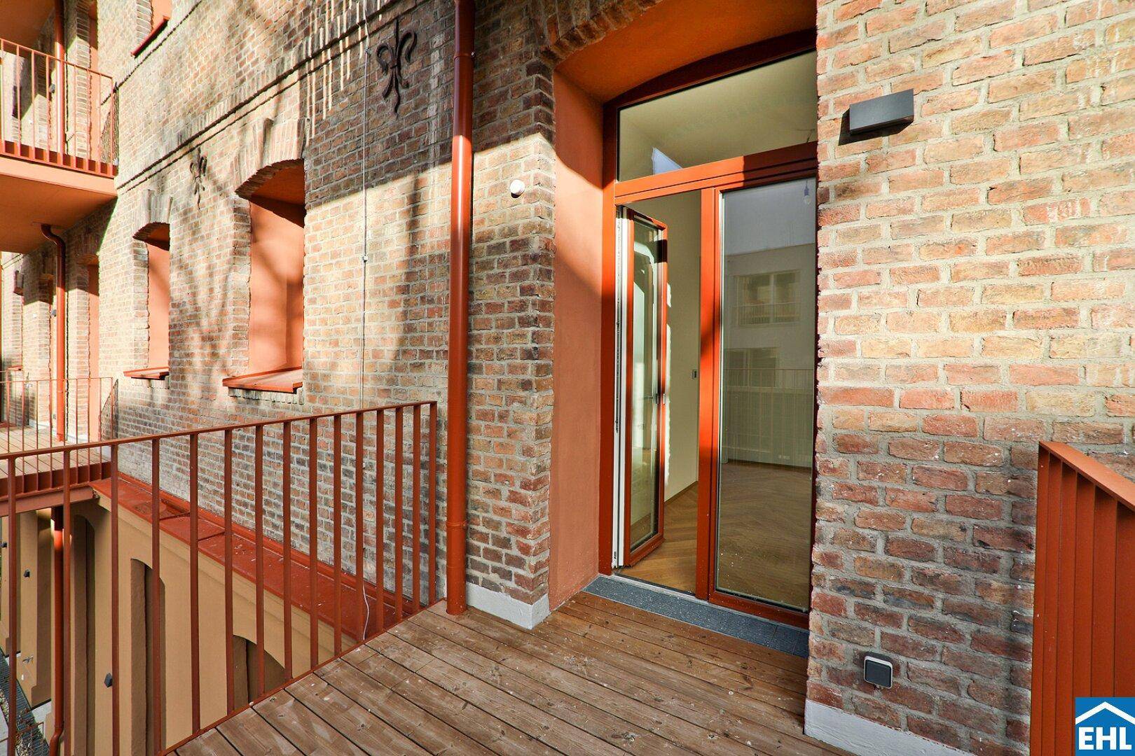 Balkon mit Holzbelag und orangefarbenem Geländer, angrenzend an eine Ziegelsteinfassade.