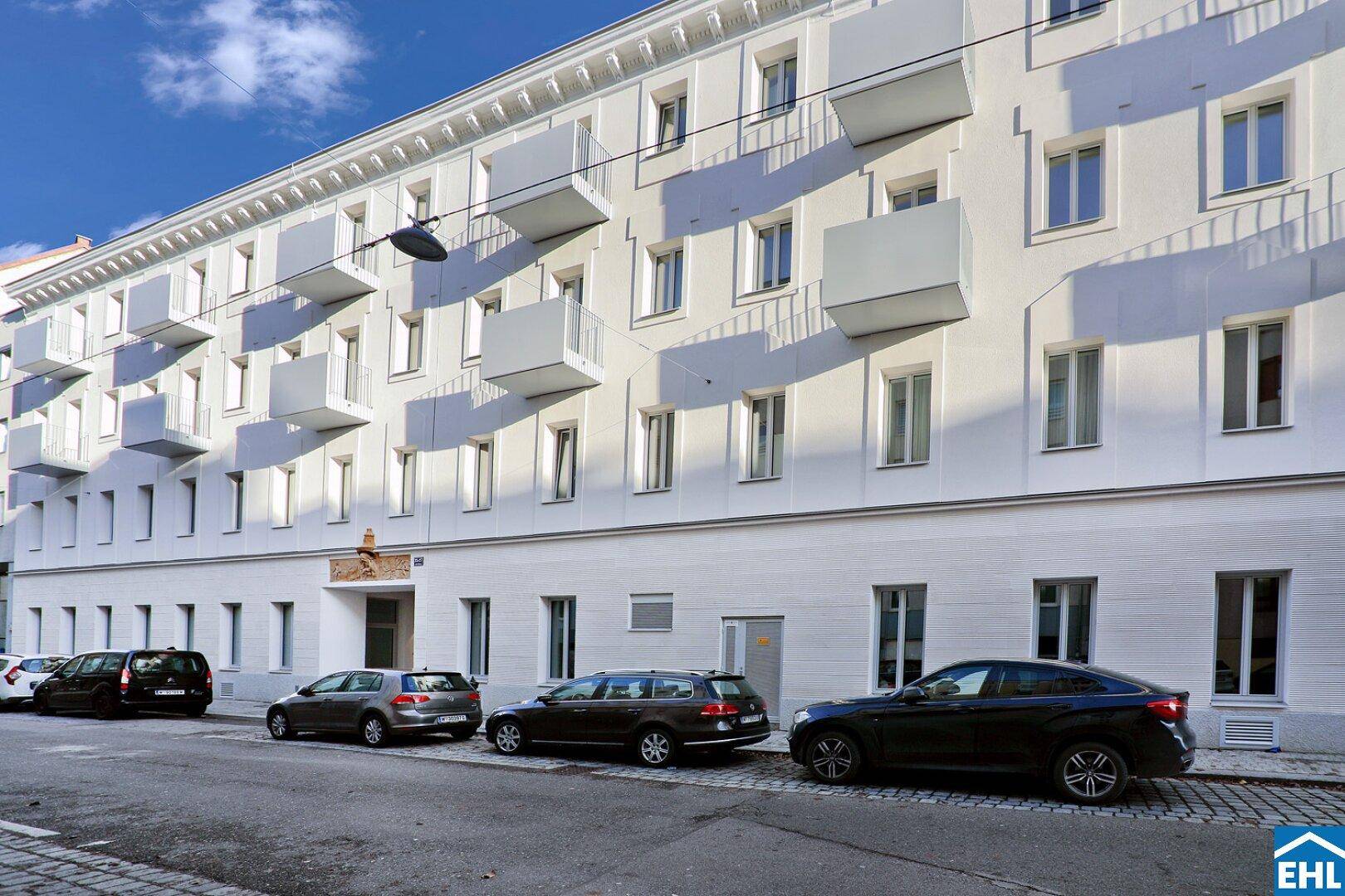 Weiße Gebäudefassade mit vielen Balkonen und geparkten Autos an einer städtischen Straße.