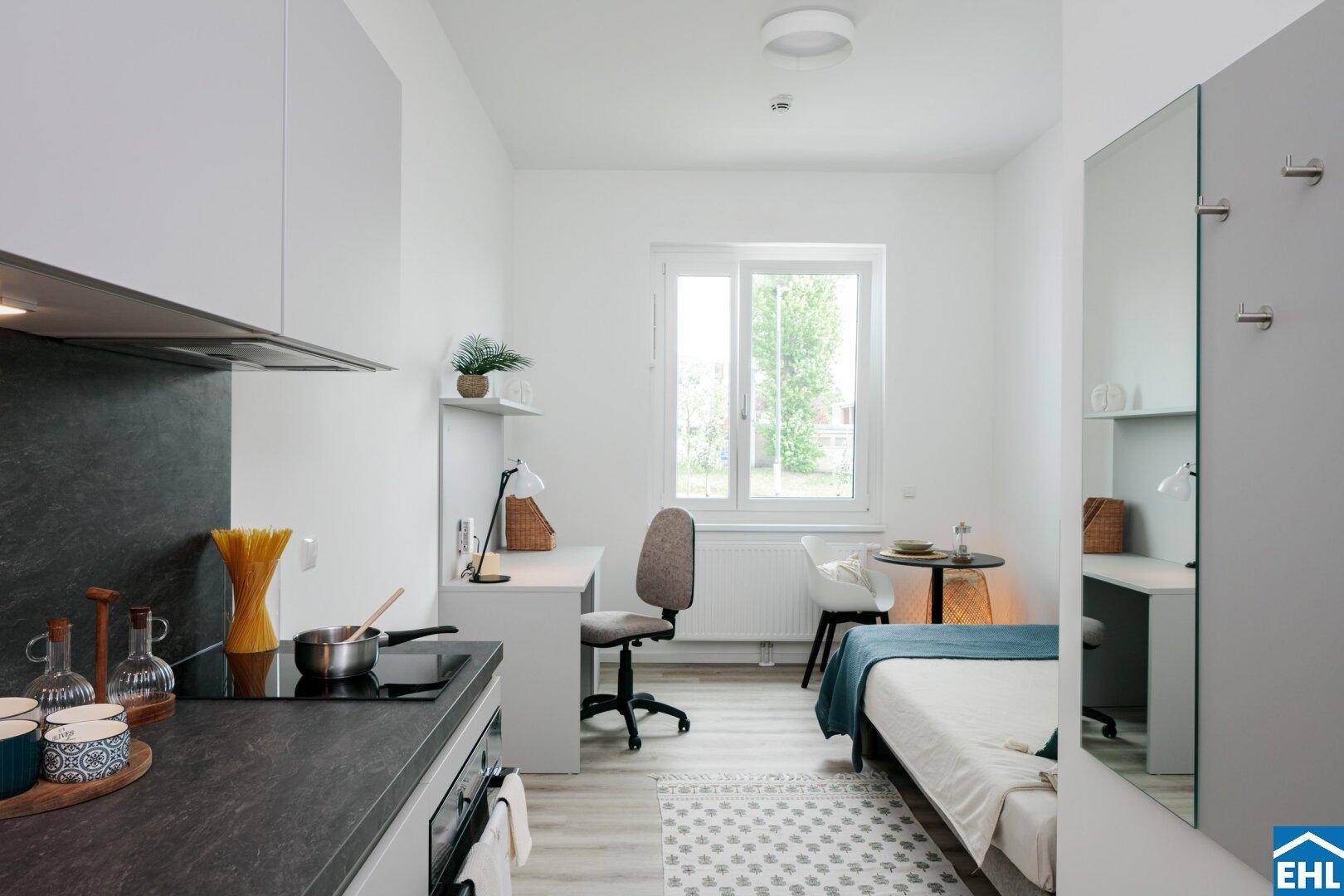 Lichtdurchflutetes Studio-Apartment mit Schreibtisch, Bett und kleiner Küchenzeile.