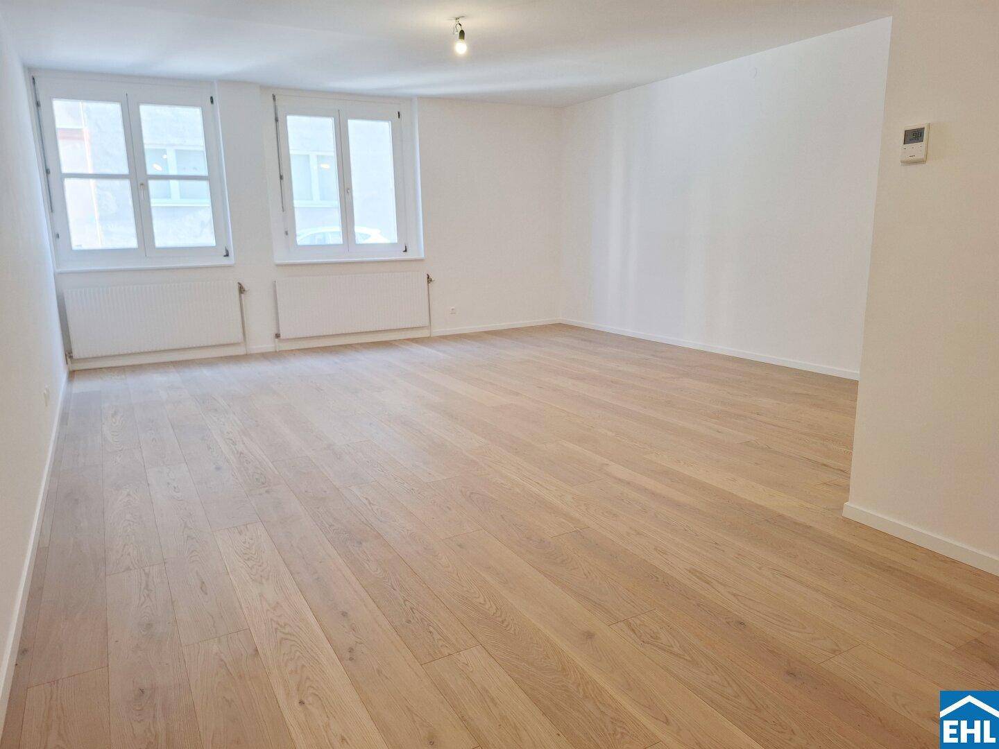 Geräumiger, leerer Wohnraum mit hellem Parkettboden und zwei großen Fenstern, bereit zur individuellen Gestaltung.