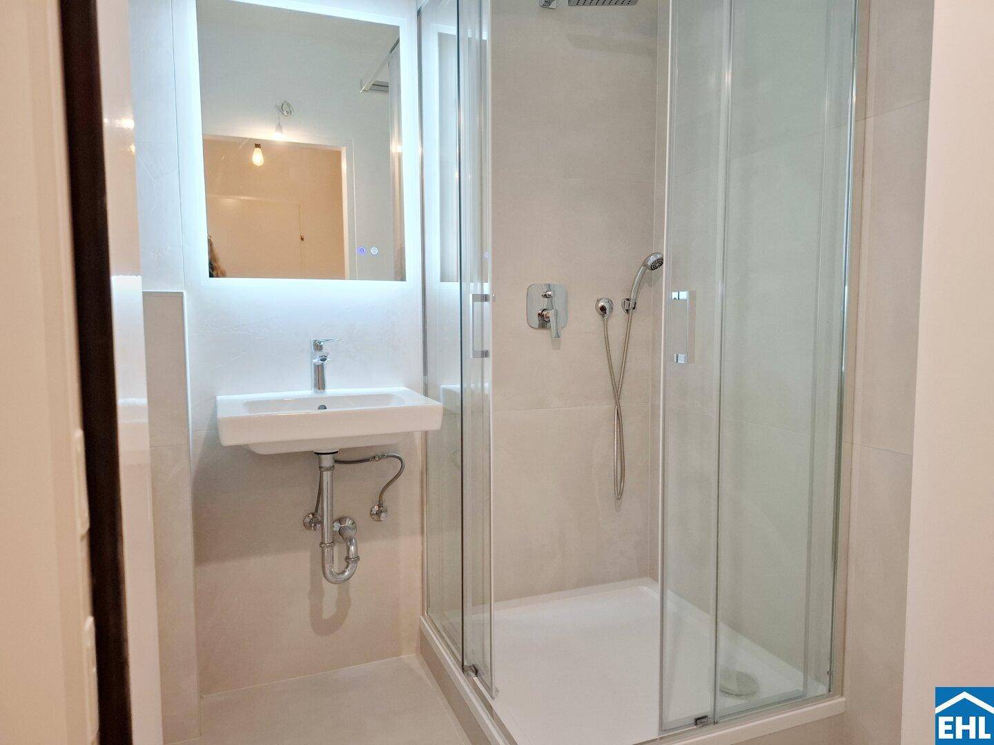 Modernes Badezimmer mit Duschkabine, beleuchtetem Spiegel und Waschbecken in minimalistischem Design.