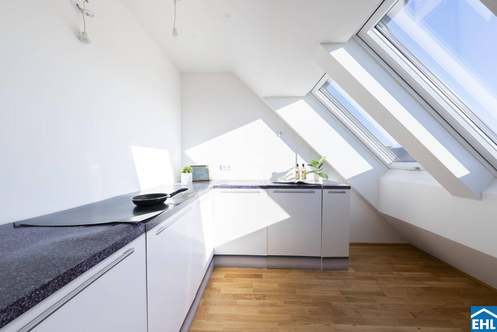 Moderne Küche mit weißen Schränken, dunkler Arbeitsplatte und viel Tageslicht durch Dachfenster.