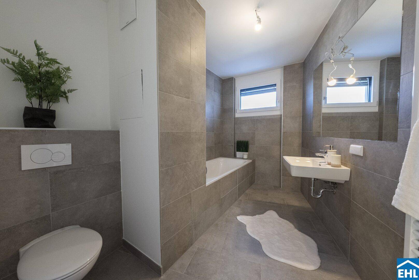 Modernes Badezimmer mit grauen Fliesen, Badewanne, WC und Fenster für Tageslicht.