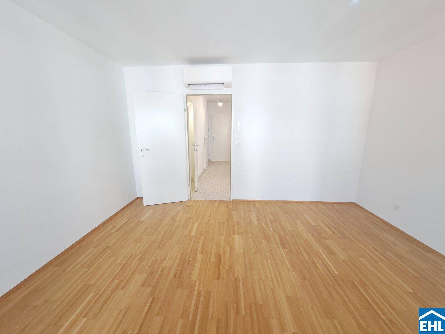 Leerer Raum mit Holzboden und Zugang zu einem weiteren Bereich, hell und neutral gestaltet.
