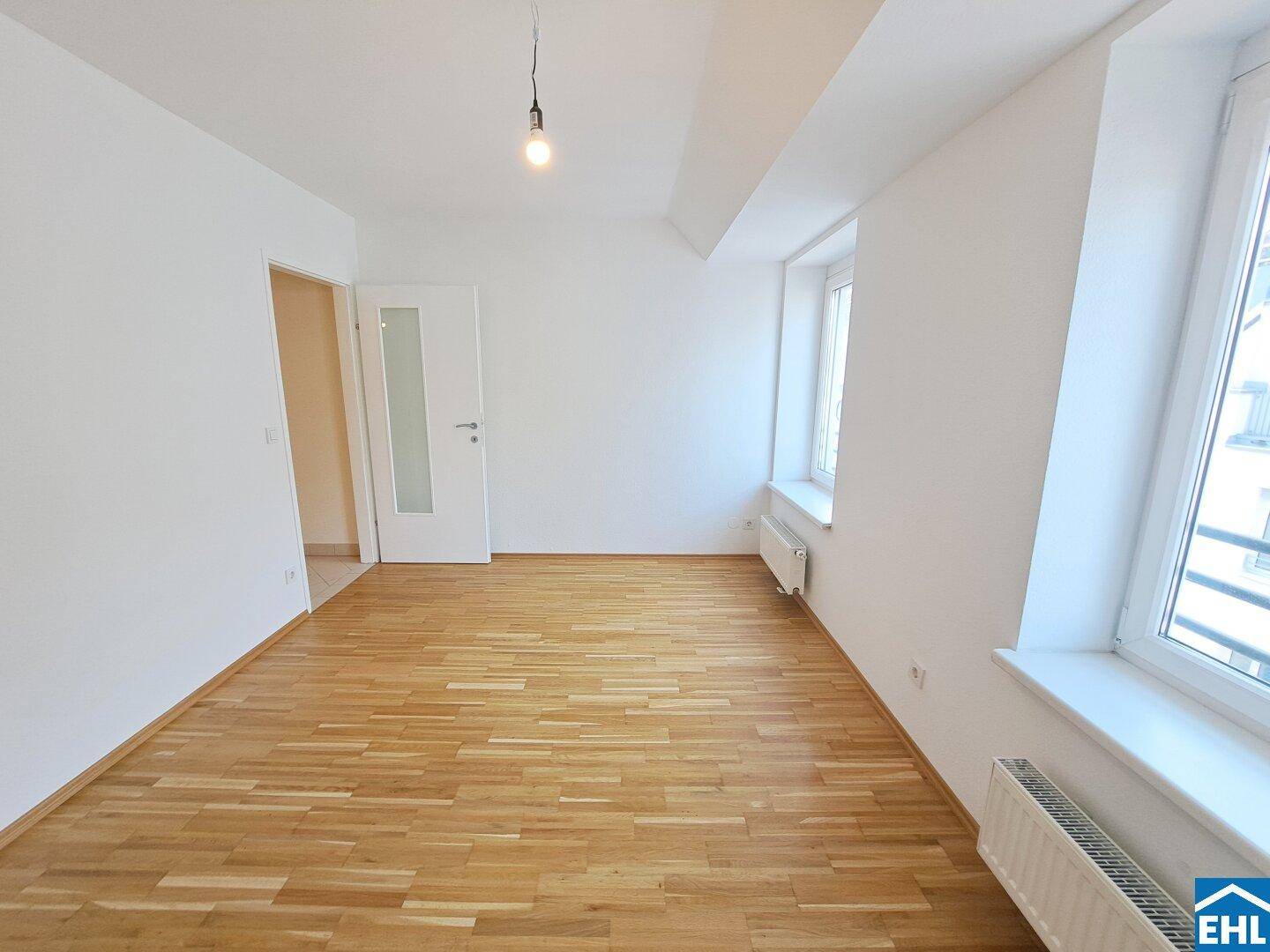 Heller Raum mit Parkettboden, weißen Wänden und großen Fenstern.