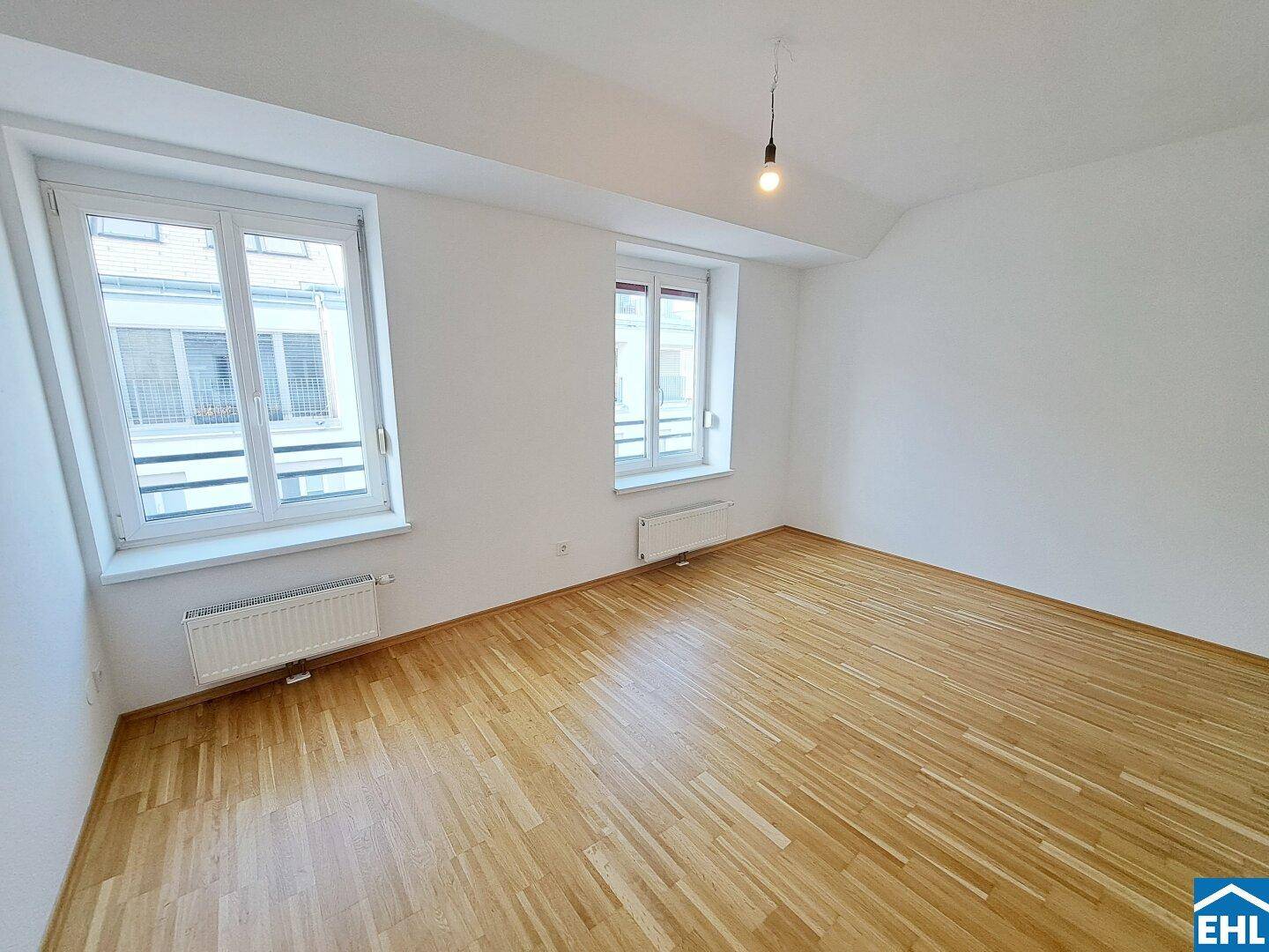 Geräumiges Zimmer mit hellem Parkettboden, weißen Wänden und zwei großen Fenstern.