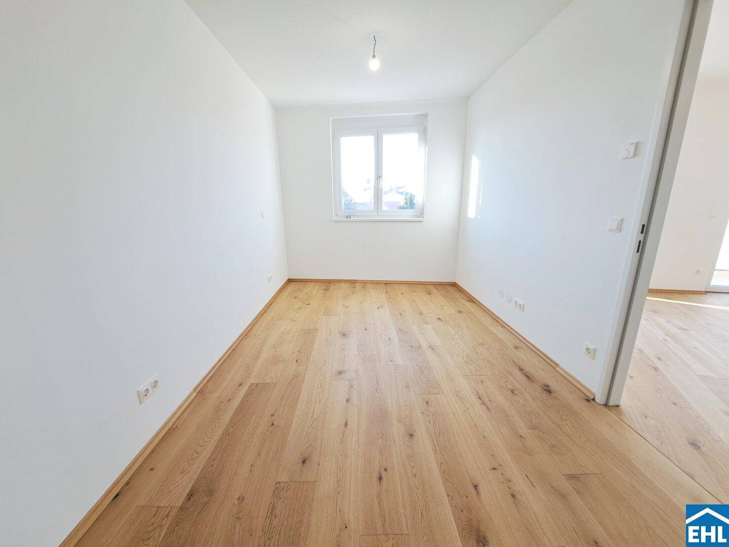 Leeres Schlafzimmer mit Parkettboden und einem Fenster, das viel Licht hereinlässt.