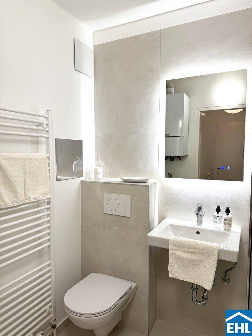 Modernes Badezimmer mit Wand-WC, beleuchtetem Spiegel und beheiztem Handtuchhalter in hellen Farbtönen.