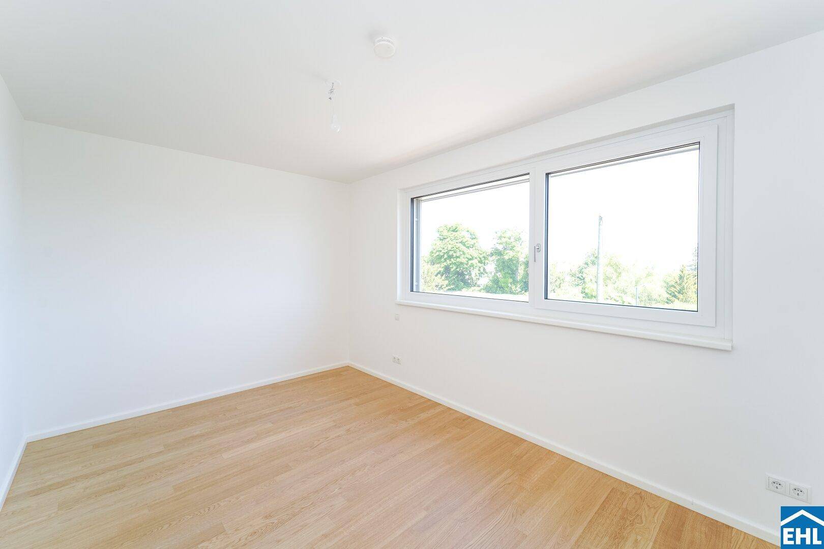 Helles Schlafzimmer mit Parkettboden, weißen Wänden und einem Fenster mit Blick ins Grüne.